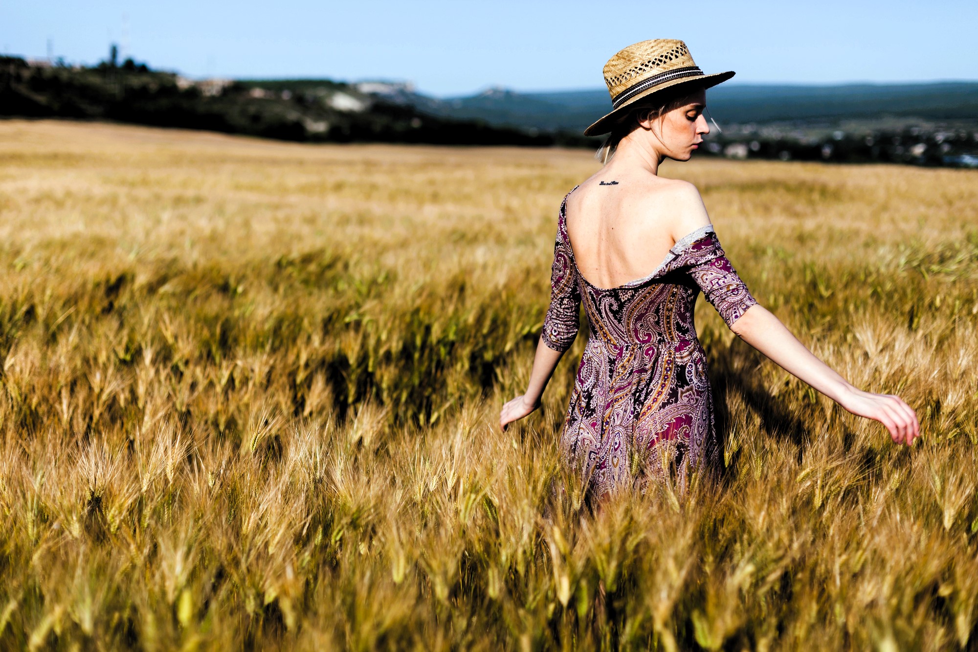 A woman in a patterned dress and straw hat walks through a golden wheat field. Her back is turned towards the camera, and the sky is clear. She gently touches the wheat stalks with her hand.