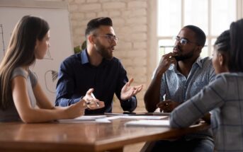 Four people are sitting around a table in an office setting, engaged in a discussion. Two men and two women are involved, with one man gesturing while speaking. A whiteboard with diagrams is visible in the background.