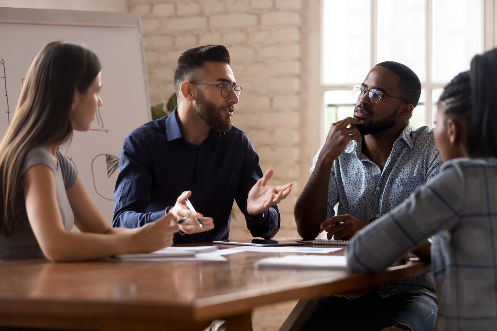 Four people are sitting around a table in an office setting, engaged in a discussion. Two men and two women are involved, with one man gesturing while speaking. A whiteboard with diagrams is visible in the background.