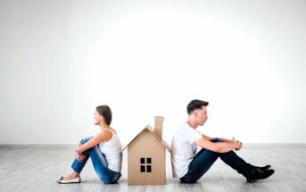 A man and a woman sit back-to-back on a light wooden floor, separated by a cardboard cutout of a house. Both are dressed casually in white shirts and jeans, looking away from each other, with the house symbolizing a barrier between them.