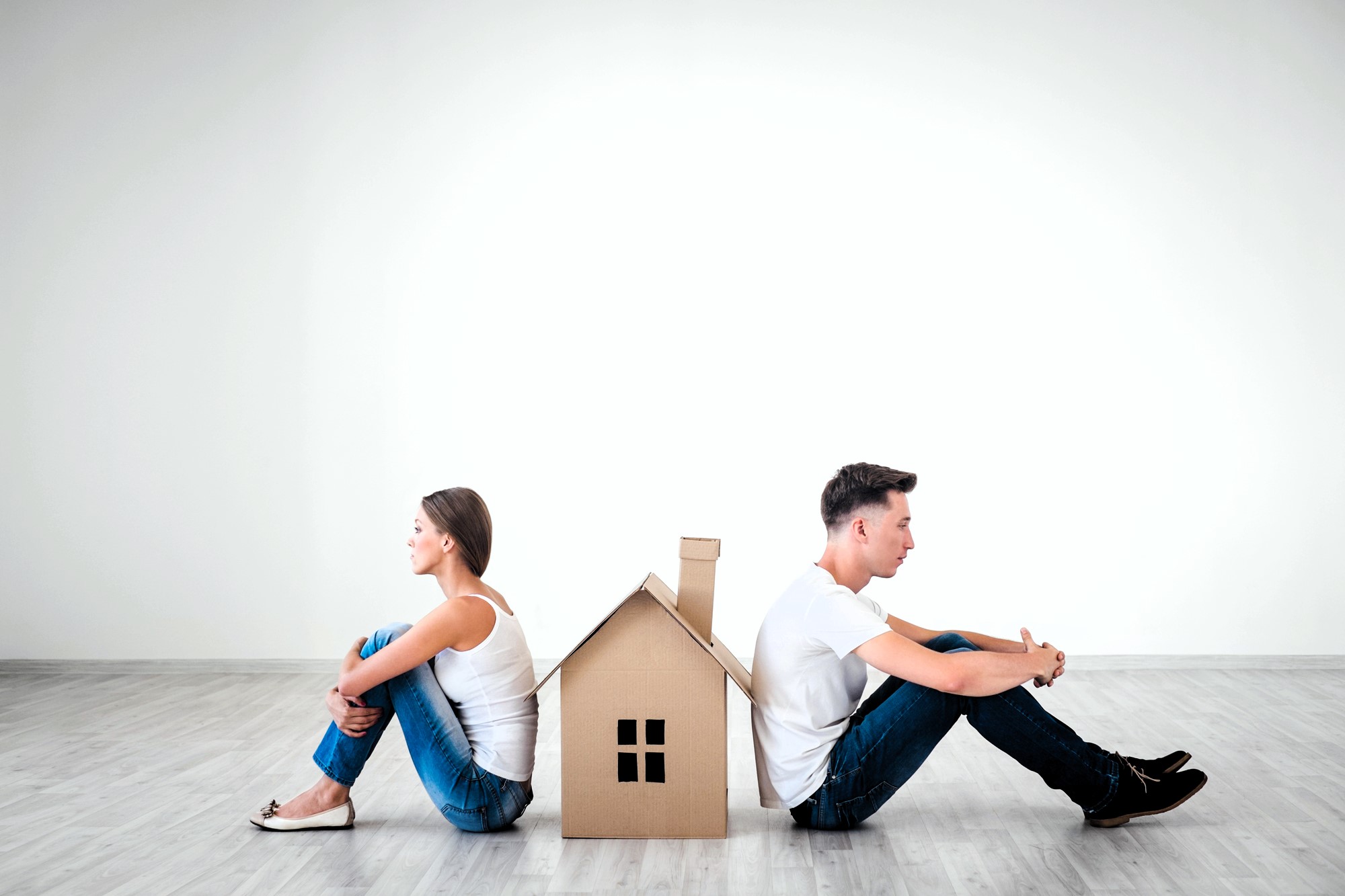 A man and a woman sit back-to-back on a light wooden floor, separated by a cardboard cutout of a house. Both are dressed casually in white shirts and jeans, looking away from each other, with the house symbolizing a barrier between them.