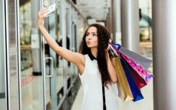 A woman with long curly hair takes a selfie while holding several colorful shopping bags in a modern mall. She wears a sleeveless blouse and stands in a corridor with glass walls and metallic columns.