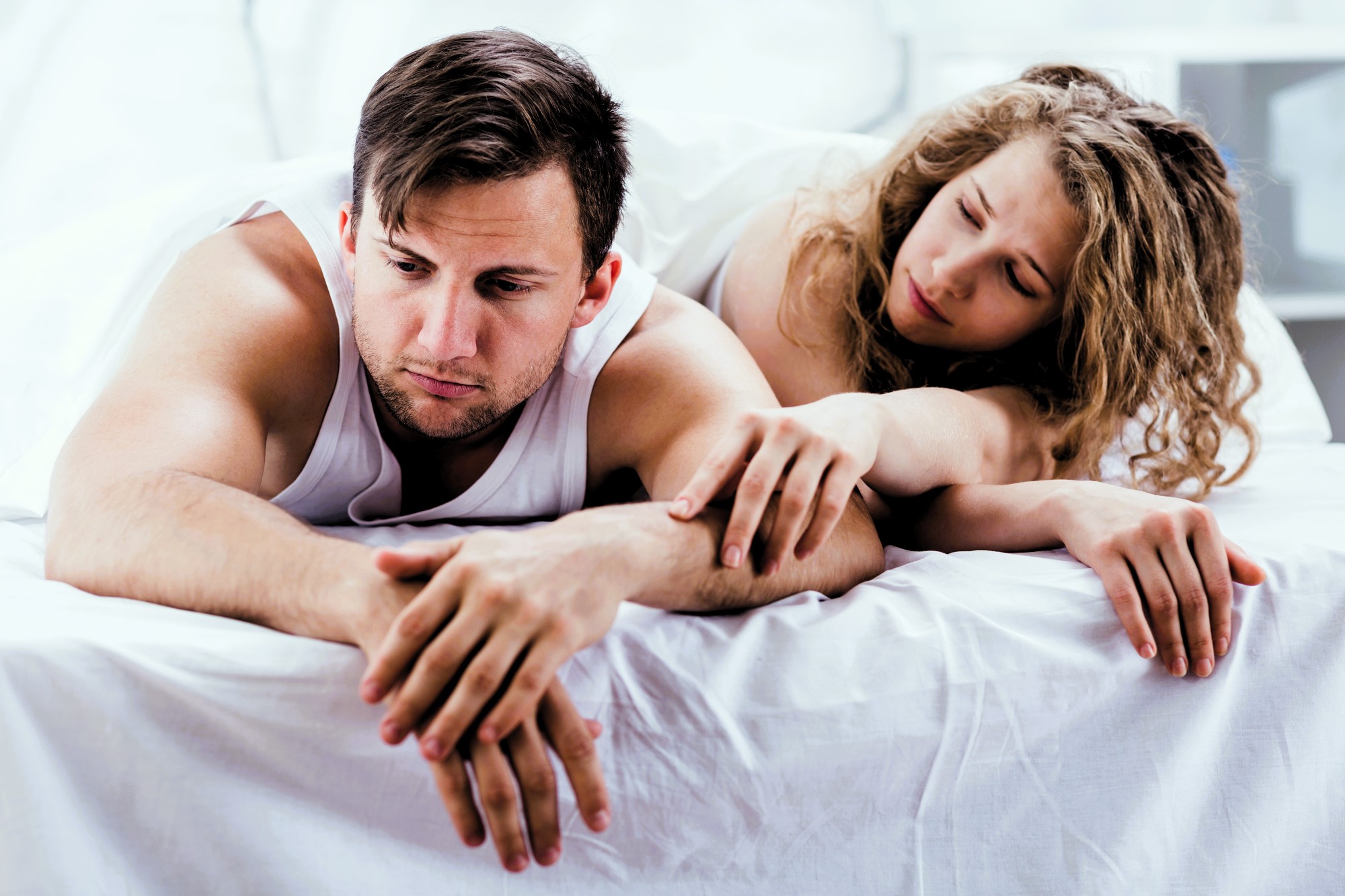 A man and a woman lie on a bed, both wearing white tank tops. The man looks upset and stares downward, while the woman gently places a hand on his arm, looking concerned. The background is softly blurred.