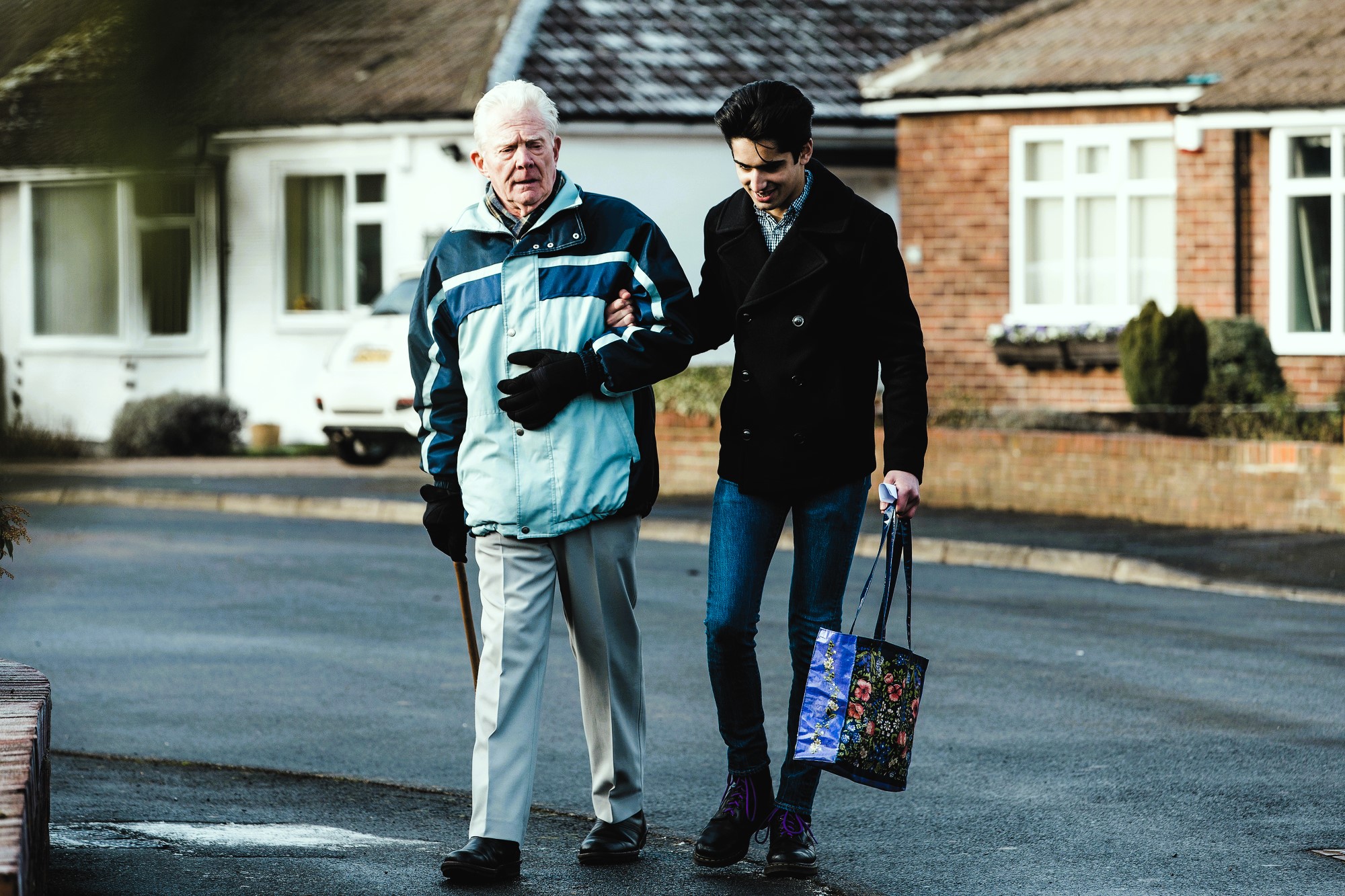 An elderly man with a cane is being helped by a younger man holding a colorful shopping bag. They are walking on a residential street with houses in the background. The older man wears a blue jacket, while the younger man is in a black coat and jeans.