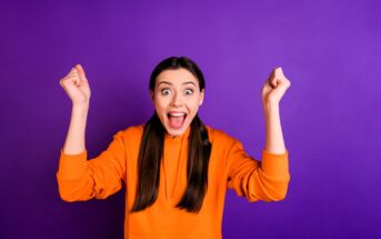 A person with long dark hair in an orange hoodie is smiling widely with their arms raised in excitement against a purple background.