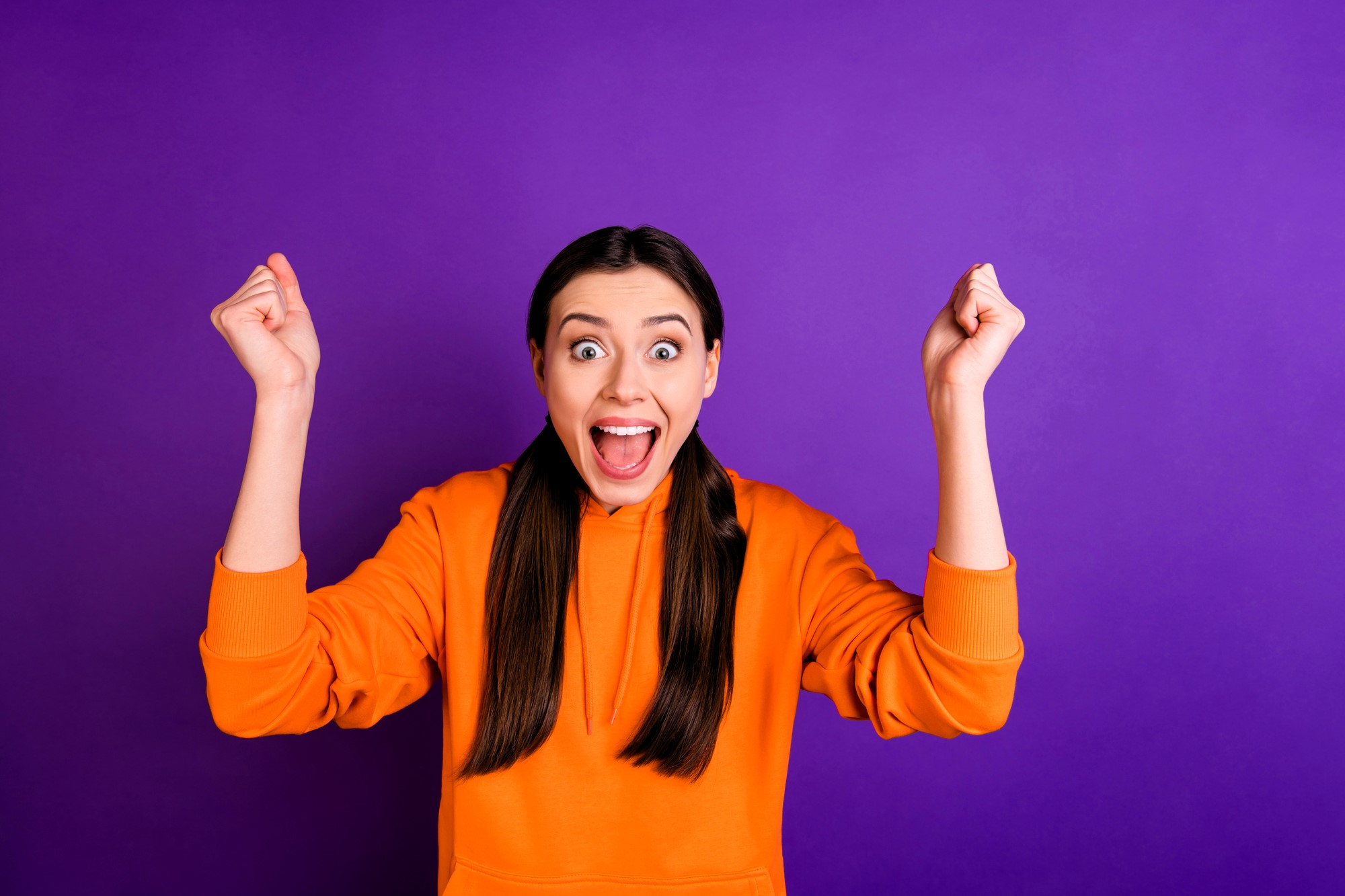 A person with long dark hair in an orange hoodie is smiling widely with their arms raised in excitement against a purple background.