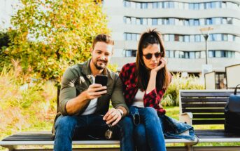 A man and woman sit on a bench outdoors. The man, in a green jacket, shows something on his phone. The woman, wearing sunglasses and a red plaid shirt, looks uninterested. They are in a park with a modern building and trees in the background.