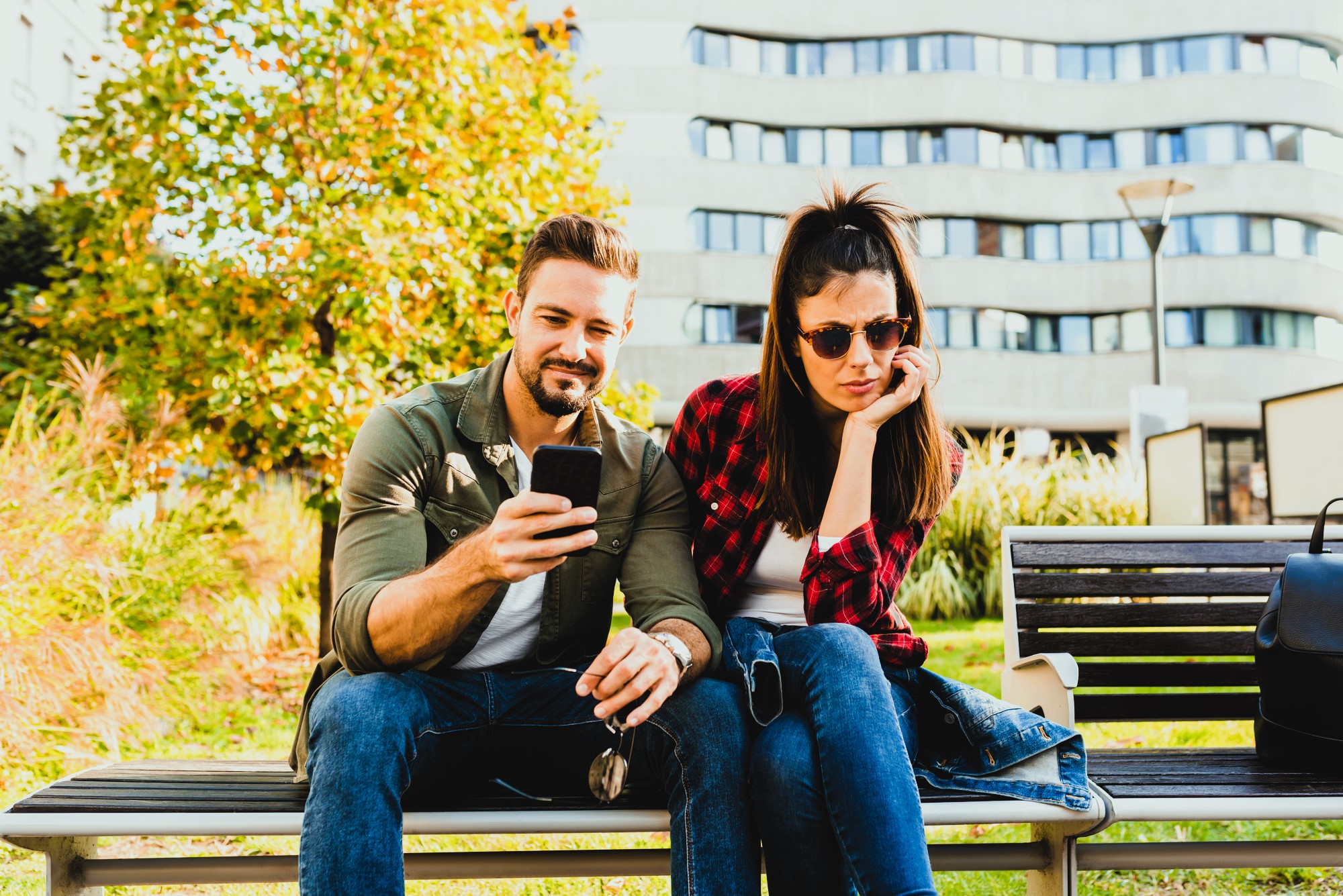 A man and woman sit on a bench outdoors. The man, in a green jacket, shows something on his phone. The woman, wearing sunglasses and a red plaid shirt, looks uninterested. They are in a park with a modern building and trees in the background.