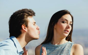 A man leans in for a kiss with puckered lips, while a woman turns her face away, appearing uninterested or reluctant. They are outdoors, with a blurred cityscape in the background.