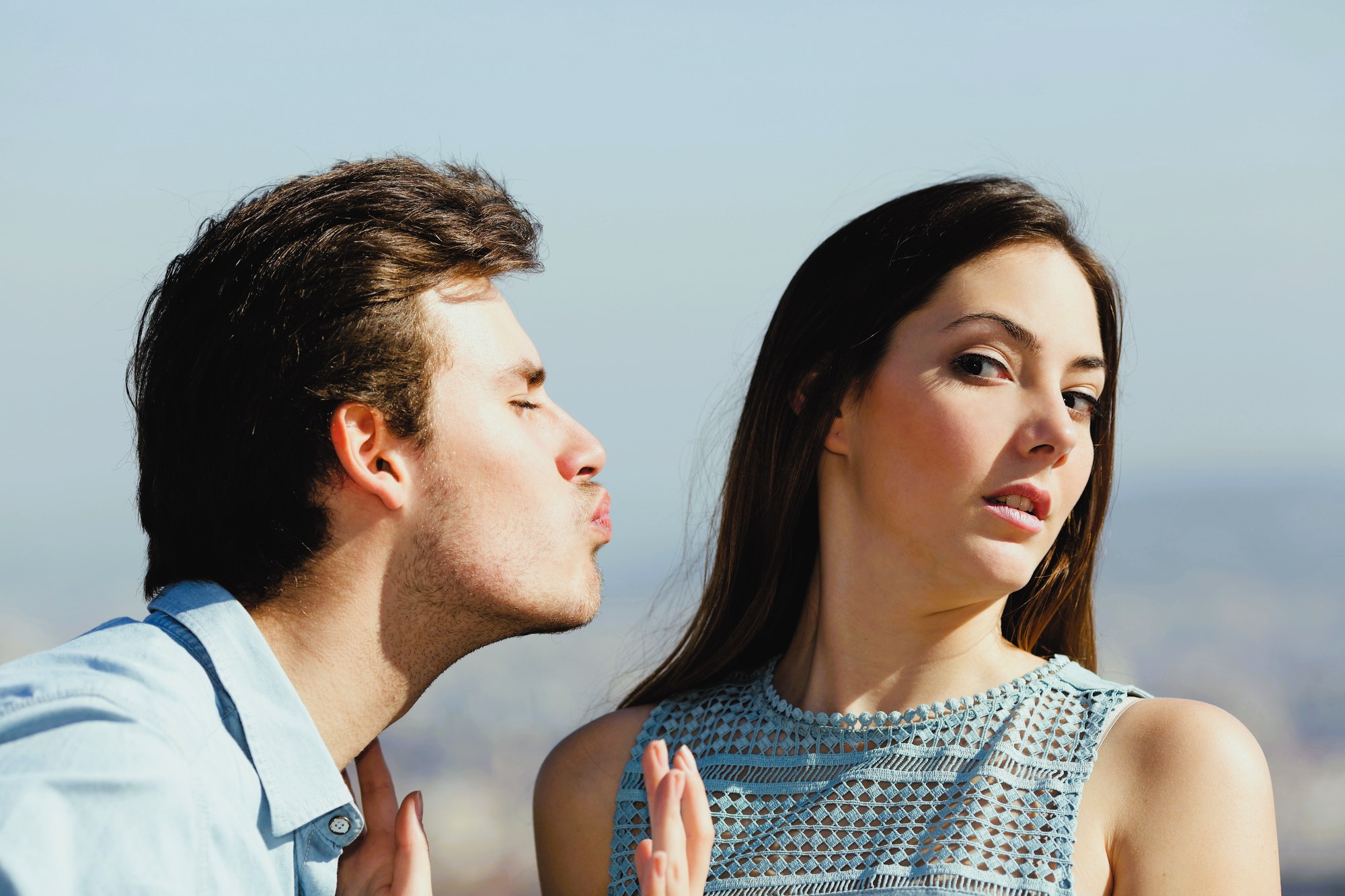 A man leans in for a kiss with puckered lips, while a woman turns her face away, appearing uninterested or reluctant. They are outdoors, with a blurred cityscape in the background.