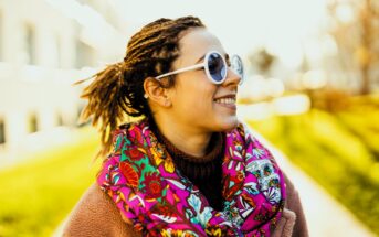 A woman wearing round sunglasses, a colorful floral scarf, and a beige coat smiles while standing outdoors. The background is a sunny park with blurred green grass and trees.