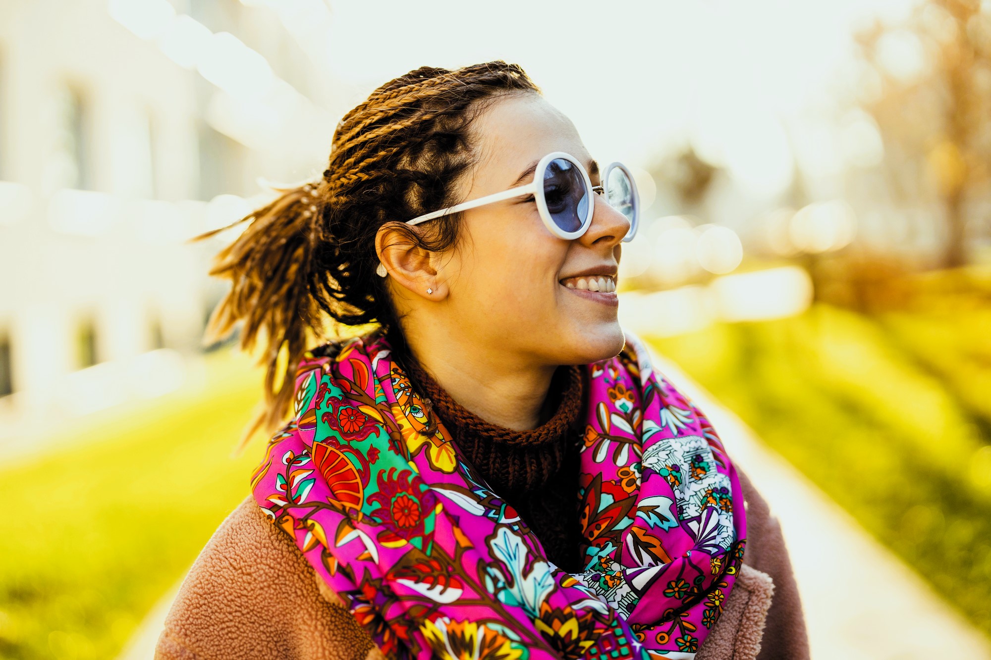 A woman wearing round sunglasses, a colorful floral scarf, and a beige coat smiles while standing outdoors. The background is a sunny park with blurred green grass and trees.