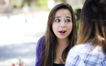 A young woman with long brown hair and expressive eyes is talking animatedly to another person outdoors. She is wearing a blue cardigan over a black top. The background is softly blurred, suggesting a park setting.