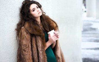 A woman with long, wavy hair is leaning against a wall while holding a coffee cup. She is wearing a brown fur coat over a green dress. The background features a snowy scene with concrete pillars.