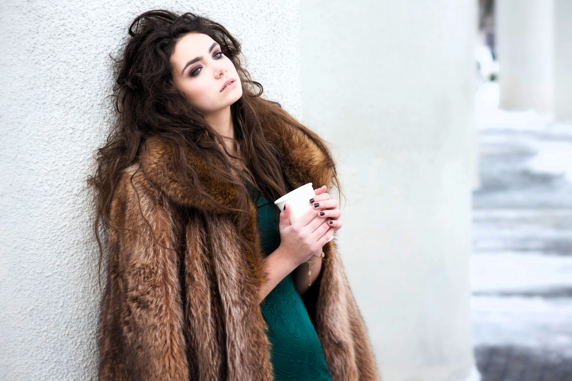 A woman with long, wavy hair is leaning against a wall while holding a coffee cup. She is wearing a brown fur coat over a green dress. The background features a snowy scene with concrete pillars.