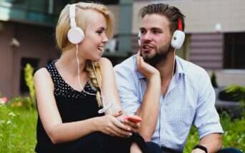 A woman and a man sit on grass, wearing headphones and smiling at each other. The woman holds a smartphone and wears a black sleeveless top. The man is in a light checkered shirt. They appear relaxed and engaged in conversation.