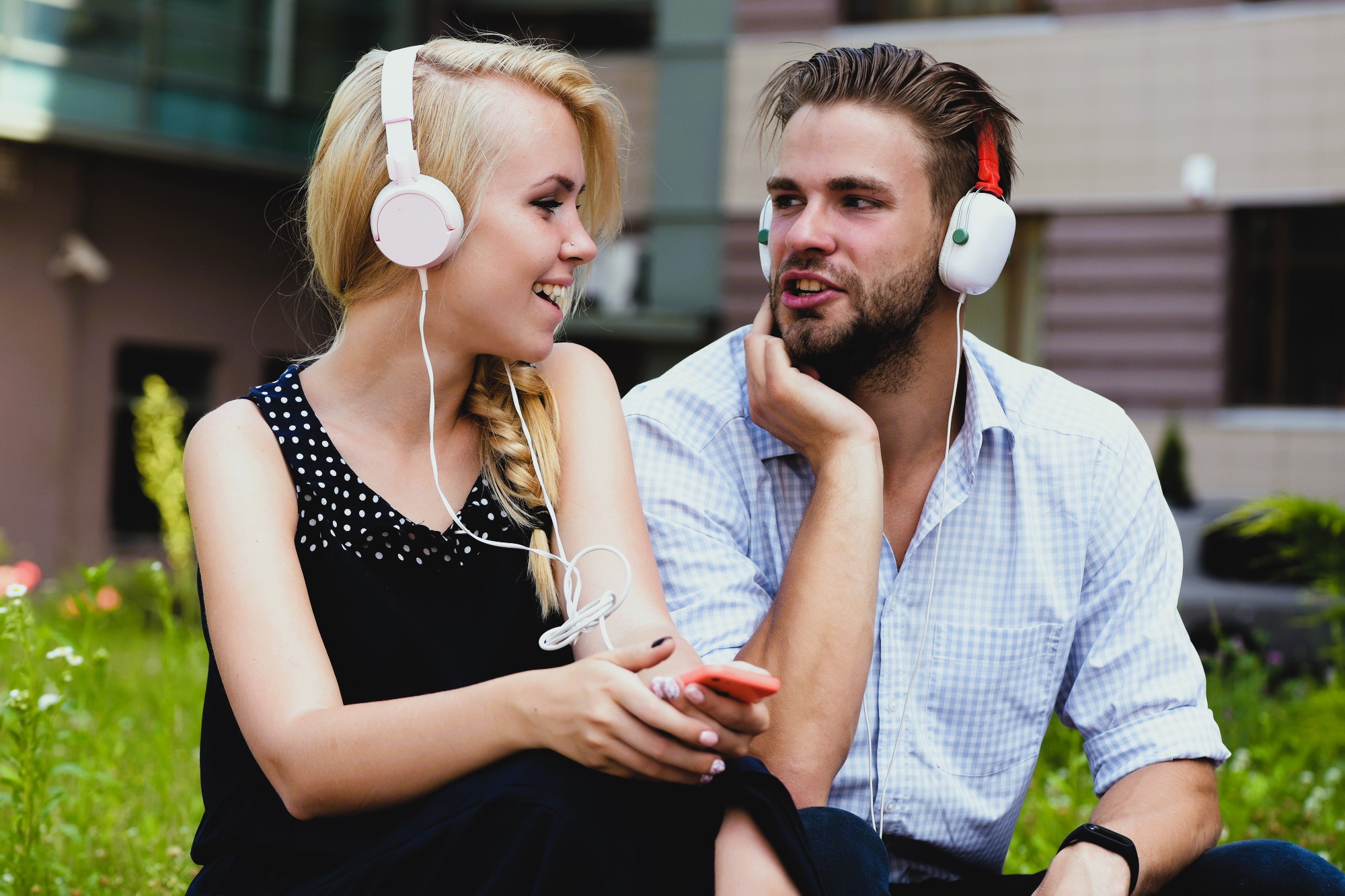 A woman and a man sit on grass, wearing headphones and smiling at each other. The woman holds a smartphone and wears a black sleeveless top. The man is in a light checkered shirt. They appear relaxed and engaged in conversation.