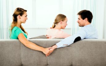 A woman and a man sit on a couch facing each other, smiling. The man is holding hands with another woman seated behind the couch, who looks concerned. The scene suggests a relationship conflict or secret encounter.