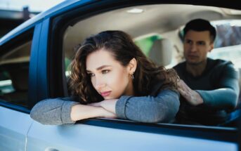 A woman with long curly hair leans on the open window of a car, appearing deep in thought. A man sits in the driver's seat, looking at her. The scene appears calm and introspective.