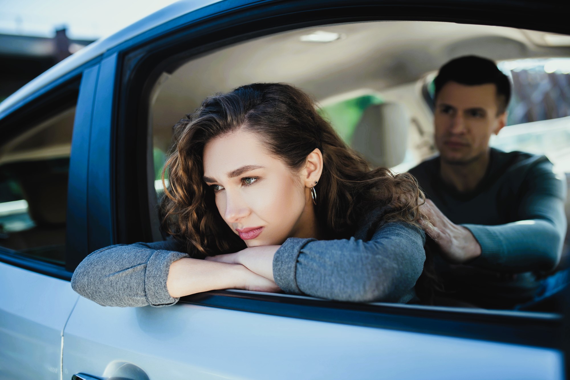 A woman with long curly hair leans on the open window of a car, appearing deep in thought. A man sits in the driver's seat, looking at her. The scene appears calm and introspective.