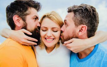 Three people cheerfully embrace. A woman in the center smiles widely, while two men on either side kiss her cheeks. They wear bright casual clothing, and the background features a clear sky.