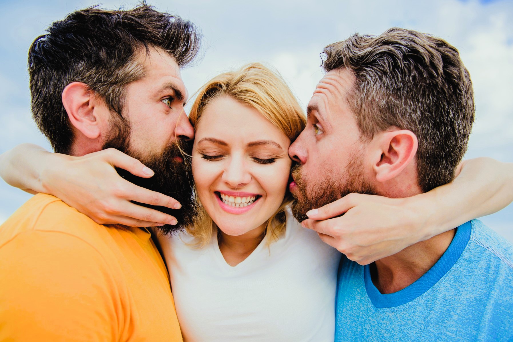 Three people cheerfully embrace. A woman in the center smiles widely, while two men on either side kiss her cheeks. They wear bright casual clothing, and the background features a clear sky.