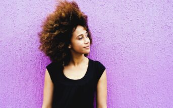 A woman with curly hair stands against a textured purple wall. She is wearing a sleeveless black top and gazes to the right with a thoughtful expression.