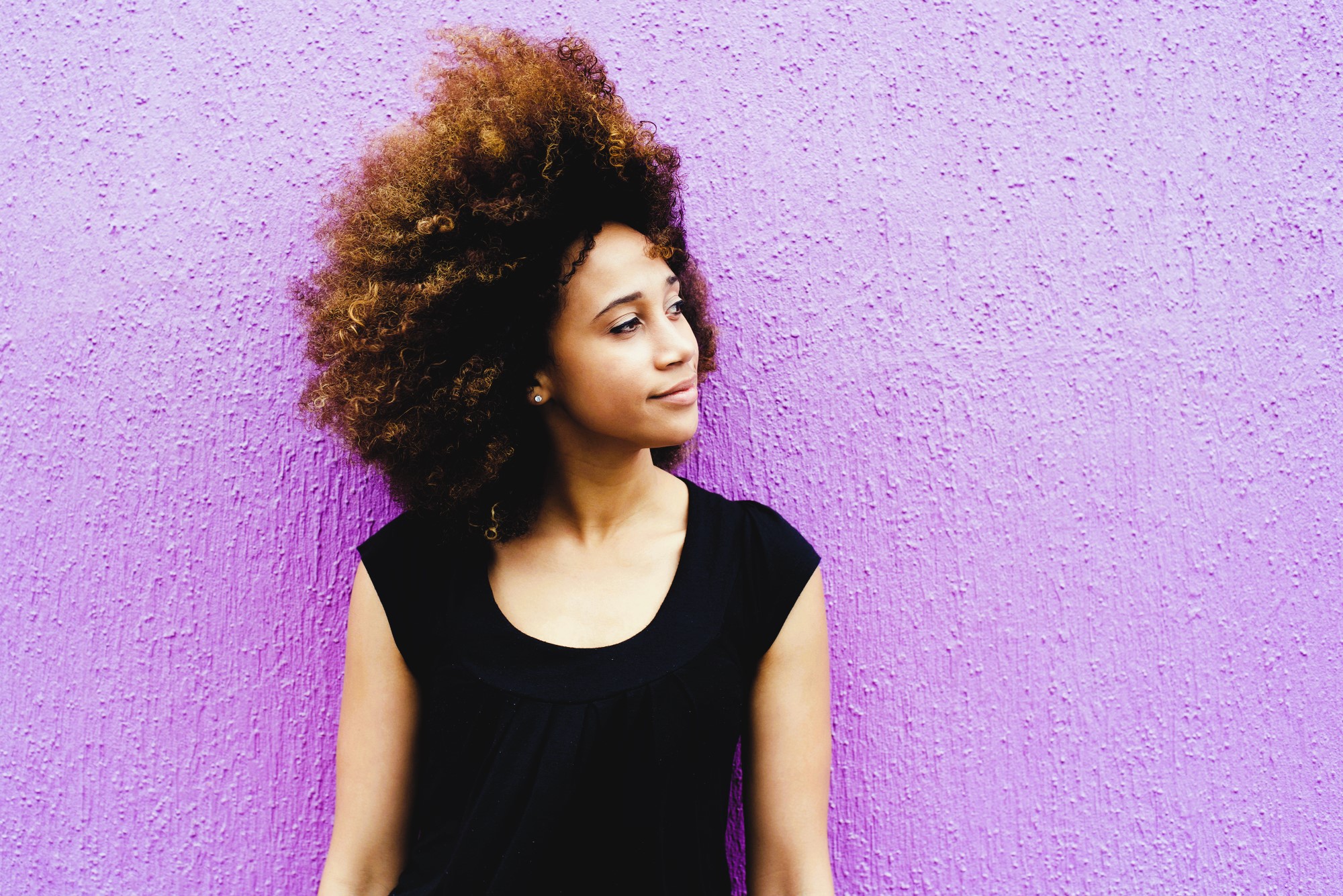 A woman with curly hair stands against a textured purple wall. She is wearing a sleeveless black top and gazes to the right with a thoughtful expression.