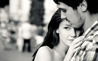 A black and white image of a young couple embracing outdoors. The woman looks at the camera with a thoughtful expression, while resting her head on the man's chest. The man gazes downward. The background is blurred with people walking.