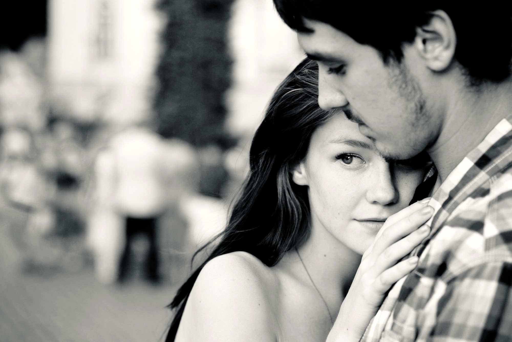 A black and white image of a young couple embracing outdoors. The woman looks at the camera with a thoughtful expression, while resting her head on the man's chest. The man gazes downward. The background is blurred with people walking.