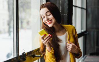 A woman with long brown hair is smiling and looking at her yellow smartphone. She is wearing a yellow cardigan over a white shirt and standing by a large window with a view of the outdoors.