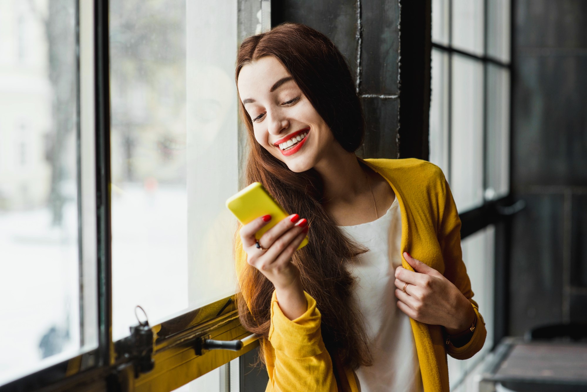 A woman with long brown hair is smiling and looking at her yellow smartphone. She is wearing a yellow cardigan over a white shirt and standing by a large window with a view of the outdoors.