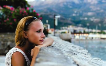 A person with short hair and a headband leans on a stone wall, gazing thoughtfully into the distance. In the background are blurred mountains, a body of water, and blooming flowers. The sky is overcast.