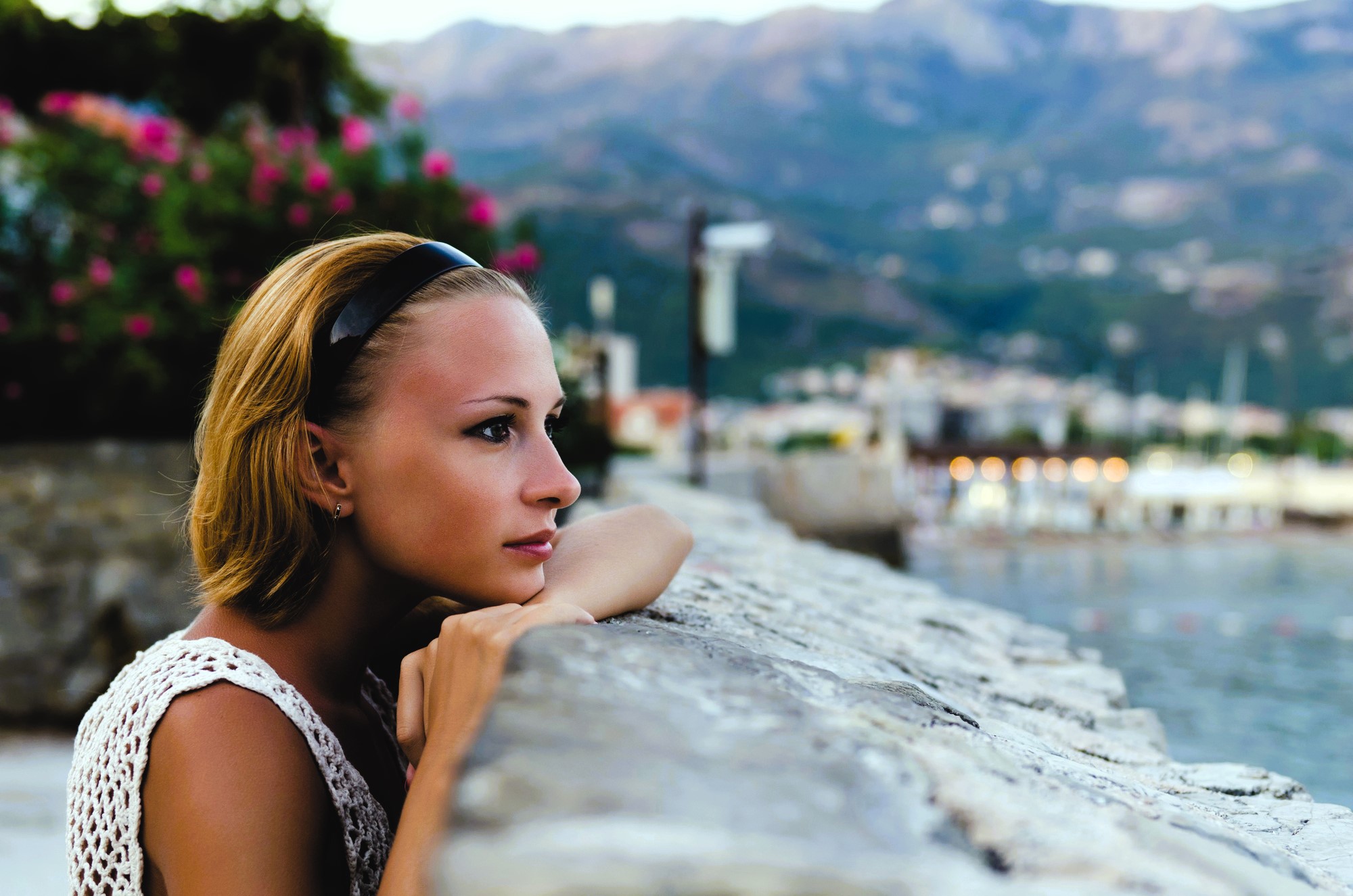 A person with short hair and a headband leans on a stone wall, gazing thoughtfully into the distance. In the background are blurred mountains, a body of water, and blooming flowers. The sky is overcast.