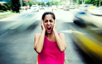 A woman in a pink top stands in the middle of a busy street, covering her ears and screaming. She appears distressed as cars blur past her, indicating motion and chaos. The background shows a cityscape with blurred vehicles.