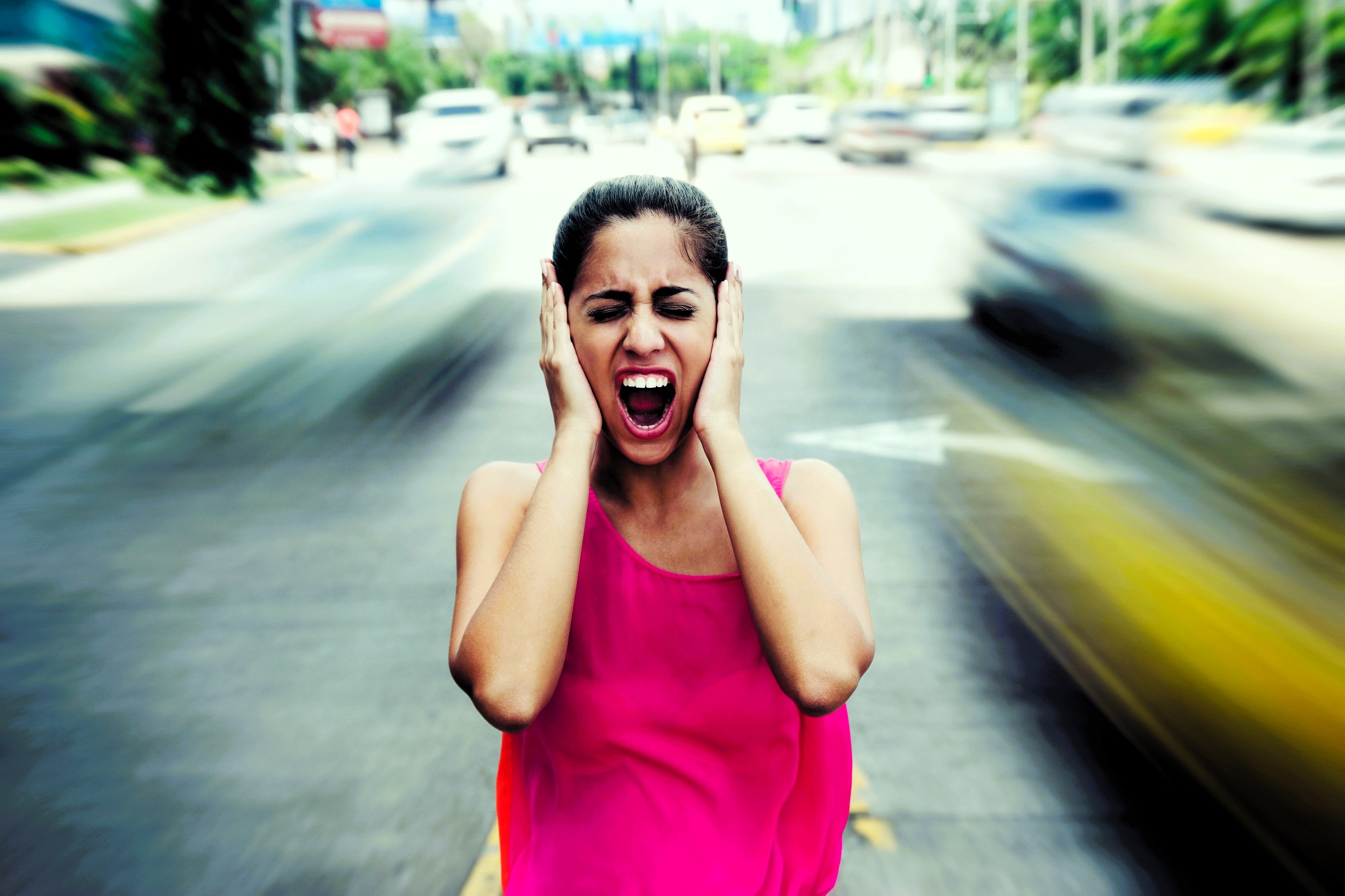 A woman in a pink top stands in the middle of a busy street, covering her ears and screaming. She appears distressed as cars blur past her, indicating motion and chaos. The background shows a cityscape with blurred vehicles.