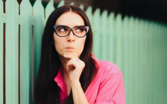 A woman with long dark hair and oversized black glasses is wearing a bright pink shirt. She stands pensively against a mint-green picket fence, resting her chin on her hand and looking to the side.