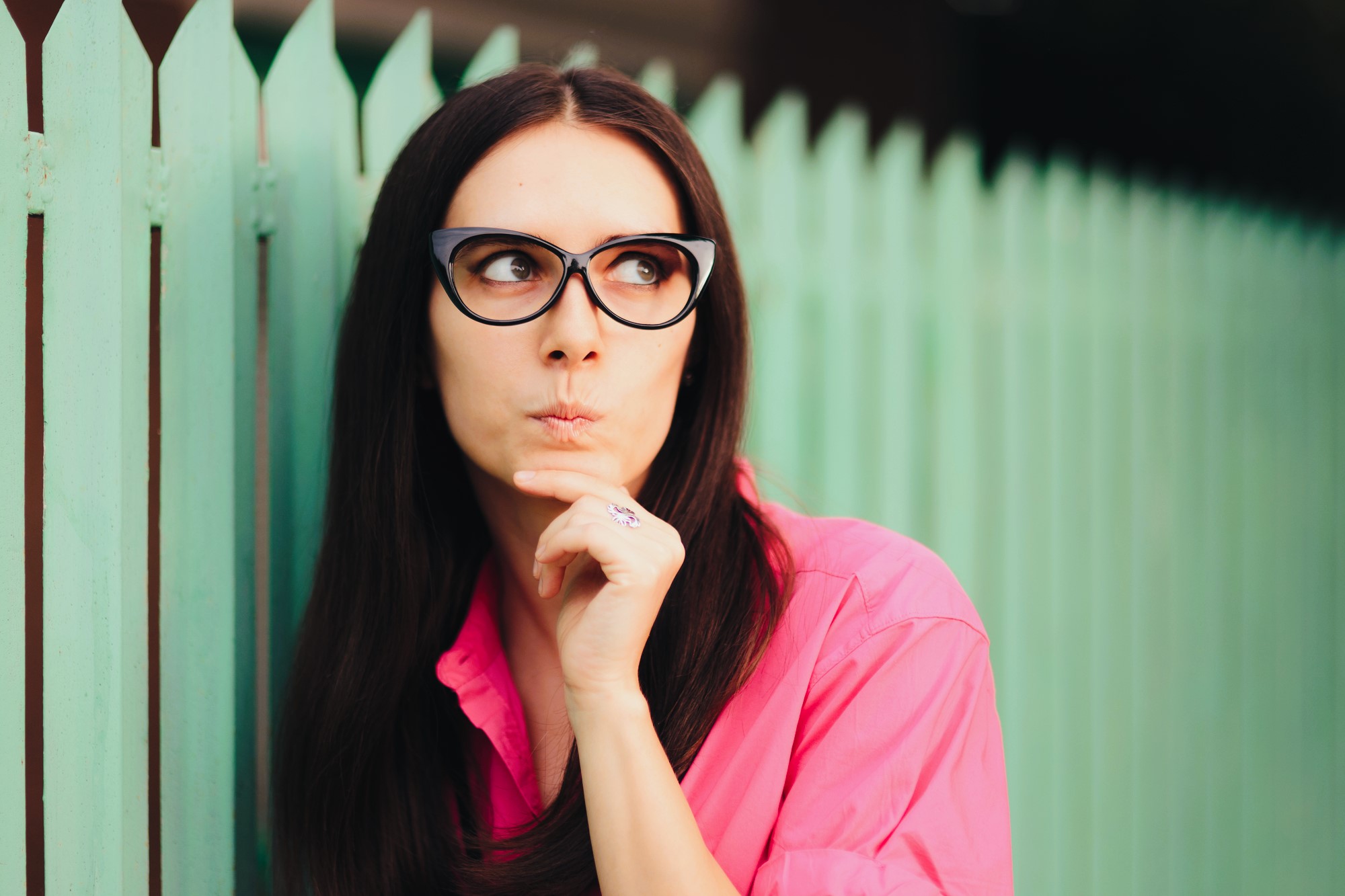 A woman with long dark hair and oversized black glasses is wearing a bright pink shirt. She stands pensively against a mint-green picket fence, resting her chin on her hand and looking to the side.