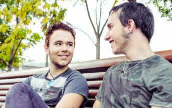 Two young men sitting on a park bench smiling at each other. They have short hair and are wearing casual t-shirts and jeans. One has earrings and a necklace. The background features trees with green and yellow leaves.