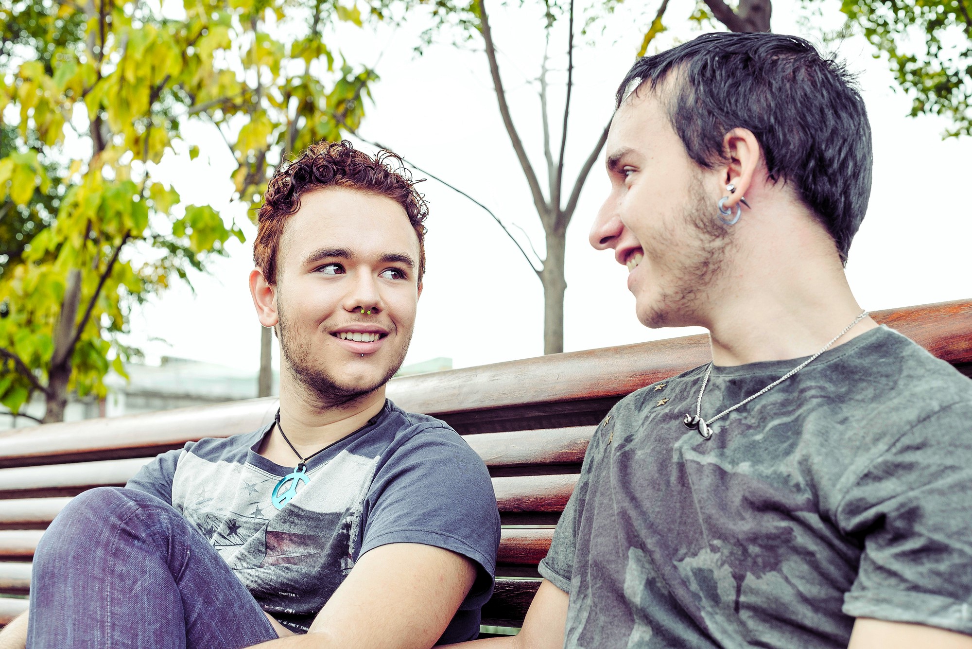 Two young men sitting on a park bench smiling at each other. They have short hair and are wearing casual t-shirts and jeans. One has earrings and a necklace. The background features trees with green and yellow leaves.