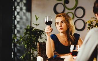 Woman sitting at a table, holding a glass of red wine, looking thoughtful. The setting appears to be a cozy restaurant with green circular wall decorations and potted plants in the background.