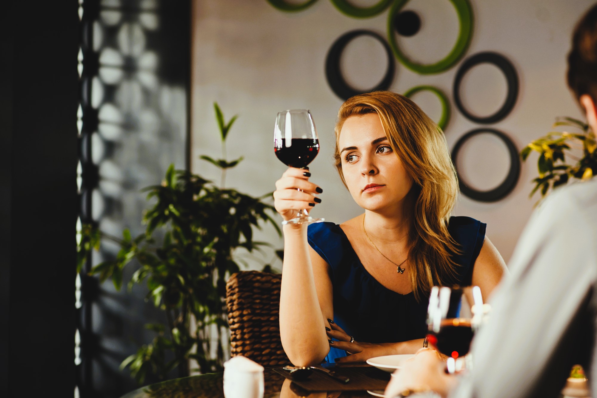 Woman sitting at a table, holding a glass of red wine, looking thoughtful. The setting appears to be a cozy restaurant with green circular wall decorations and potted plants in the background.