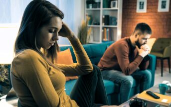 A woman and a man sitting on a couch in a living room, both appear upset and thoughtful. The woman is in the foreground with her hand on her head, while the man is in the background, leaning forward with hands clasped.