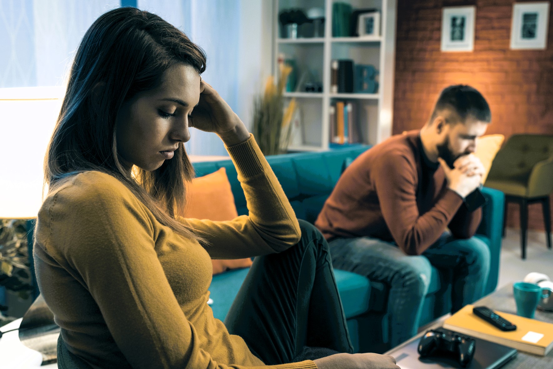 A woman and a man sitting on a couch in a living room, both appear upset and thoughtful. The woman is in the foreground with her hand on her head, while the man is in the background, leaning forward with hands clasped.