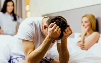 A man sits on a bed with his head in his hands, appearing distressed. A woman sits on the bed behind him, looking concerned with her hand on her mouth. Another woman stands in the background with an upset expression.