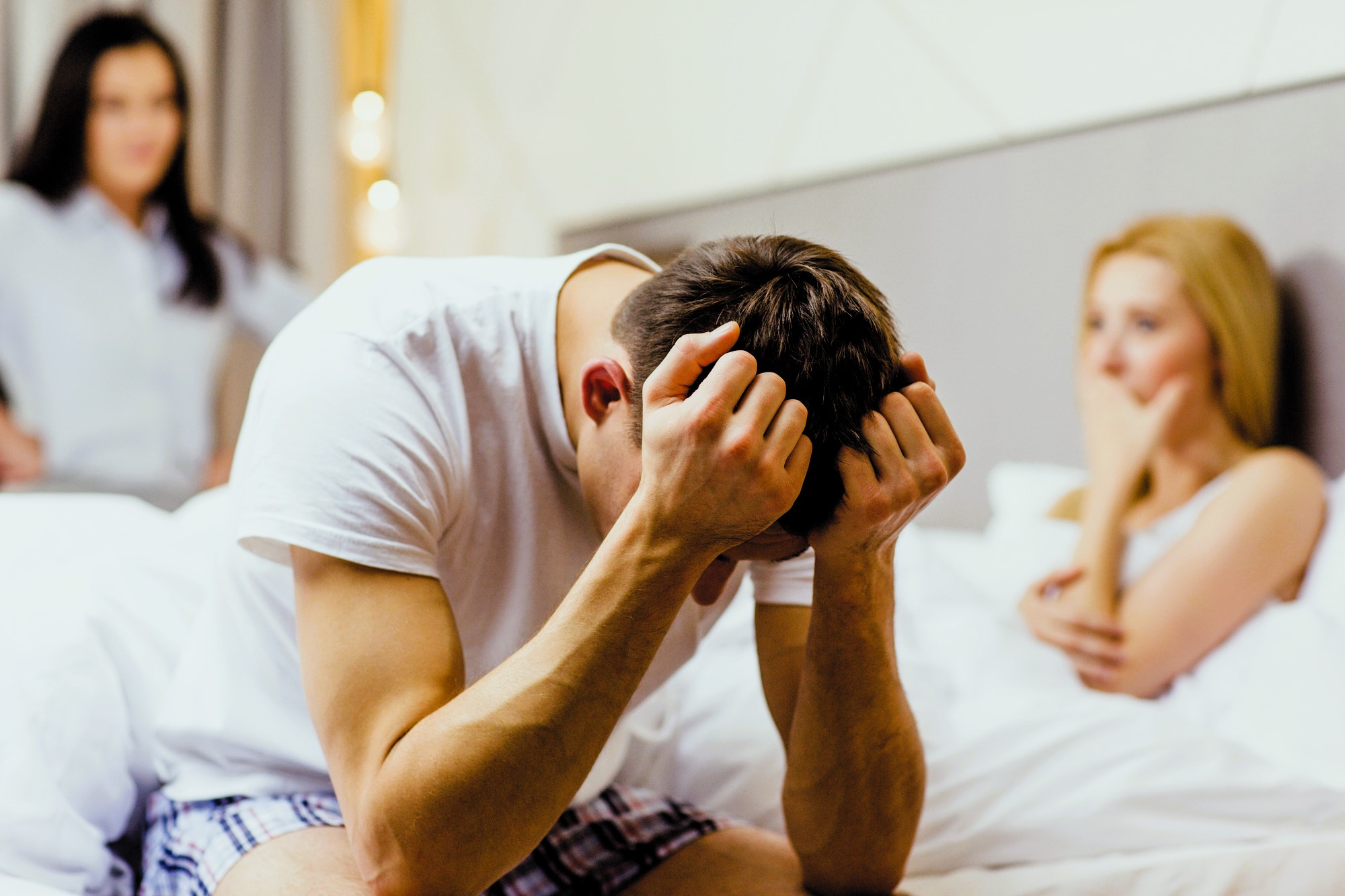 A man sits on a bed with his head in his hands, appearing distressed. A woman sits on the bed behind him, looking concerned with her hand on her mouth. Another woman stands in the background with an upset expression.