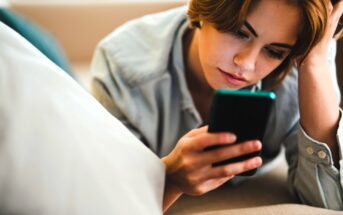 A person with short brown hair is lying on a sofa, looking at a smartphone. They appear focused, resting their head on one hand. The background is softly blurred, and the person's shirt is a light blue denim.