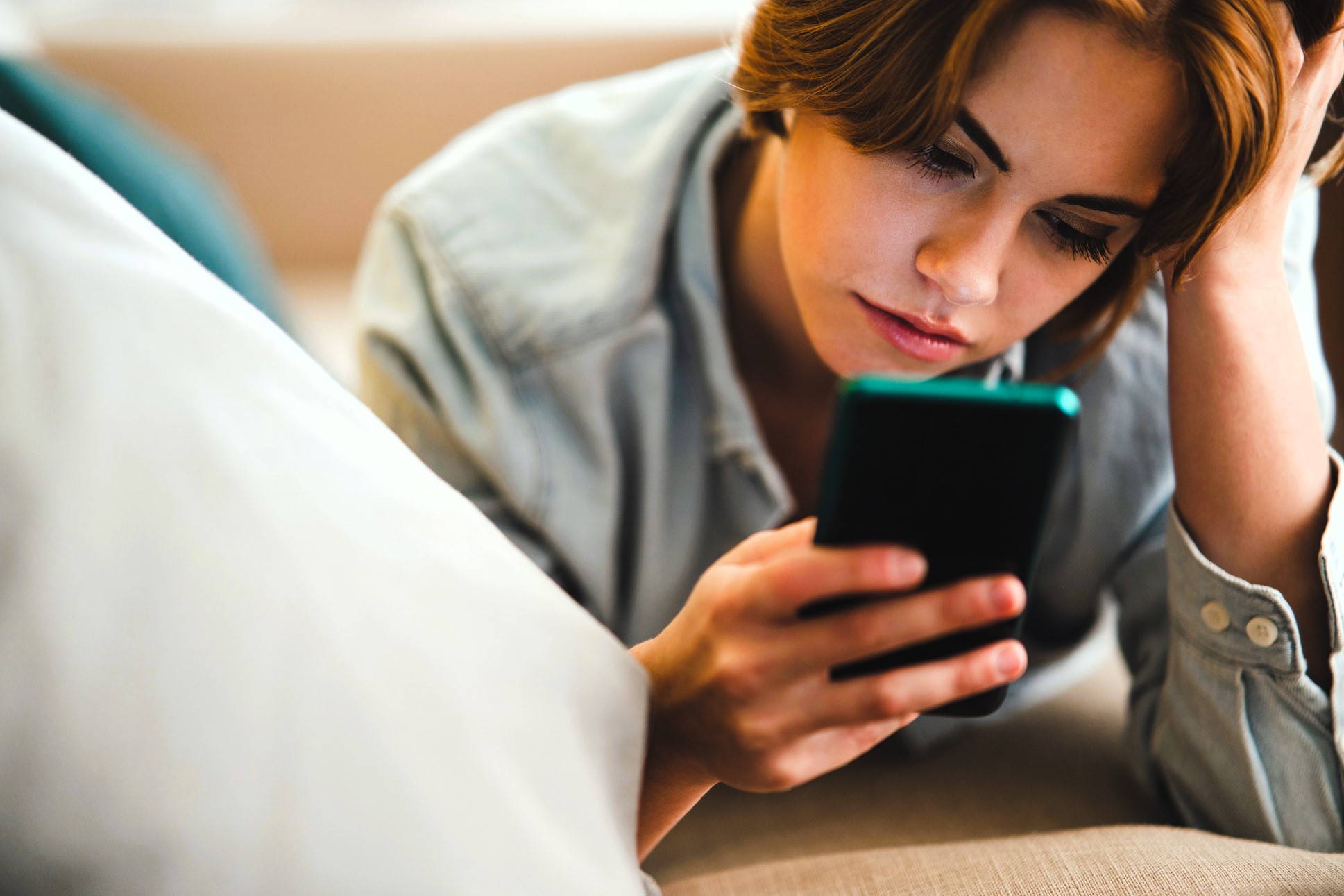 A person with short brown hair is lying on a sofa, looking at a smartphone. They appear focused, resting their head on one hand. The background is softly blurred, and the person's shirt is a light blue denim.