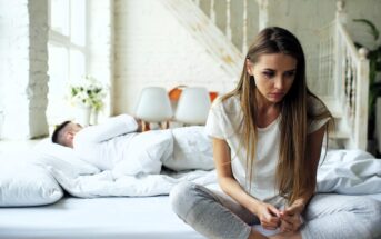 A woman sits on the edge of a bed looking thoughtful or upset, with her head slightly bowed. She is wearing a white shirt and light pants. In the background, a man lies sleeping in bed, covered with white bedding. The room has soft lighting and white decor.