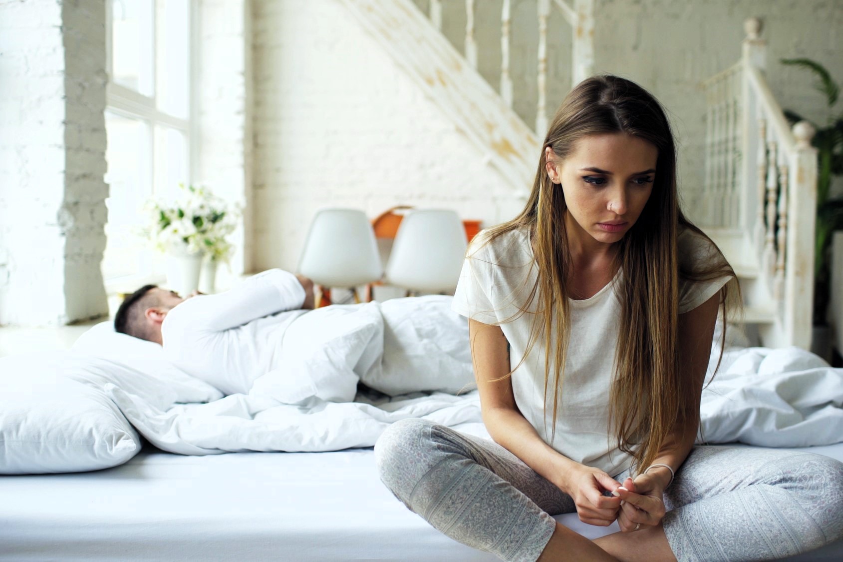 A woman sits on the edge of a bed looking thoughtful or upset, with her head slightly bowed. She is wearing a white shirt and light pants. In the background, a man lies sleeping in bed, covered with white bedding. The room has soft lighting and white decor.